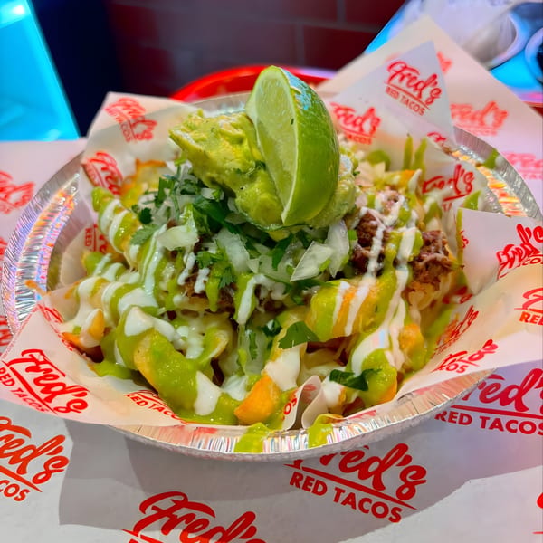 Fred's Red Tacos' loaded fries with guacamole, lime, and green and white sauces in a foil container.