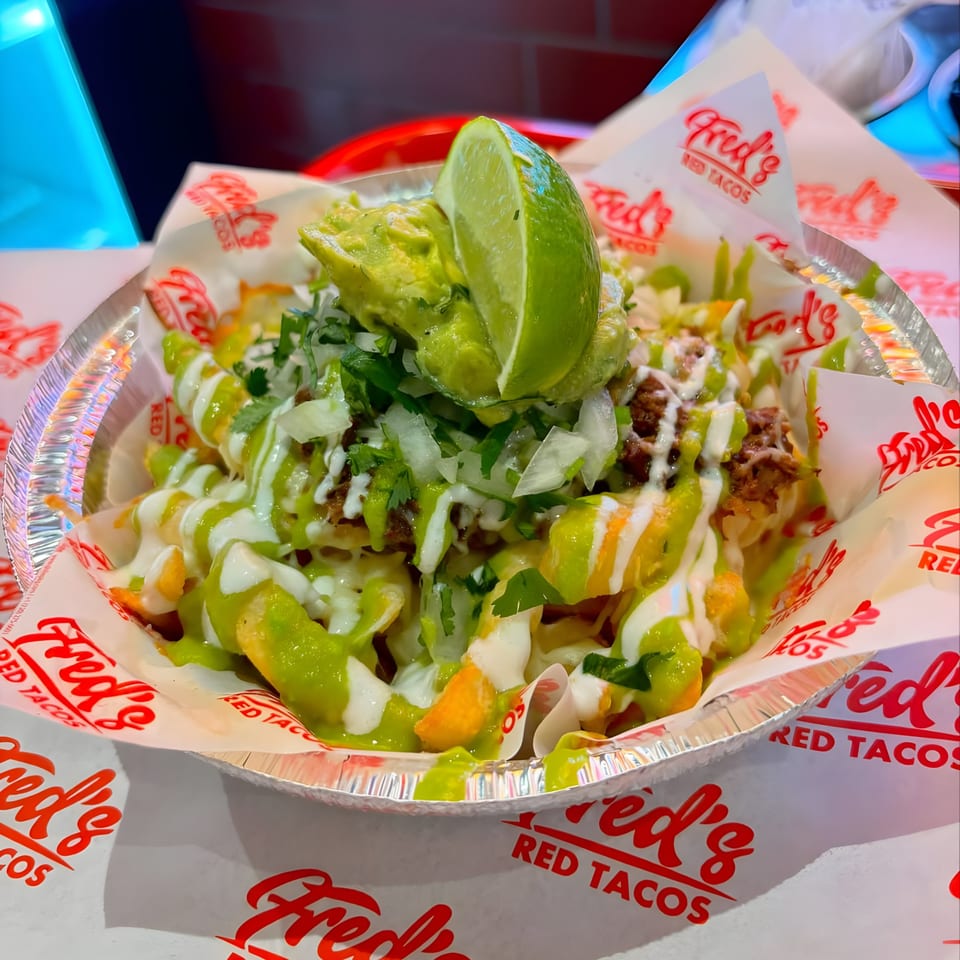 Fred's Red Tacos' loaded fries with guacamole, lime, and green and white sauces in a foil container.