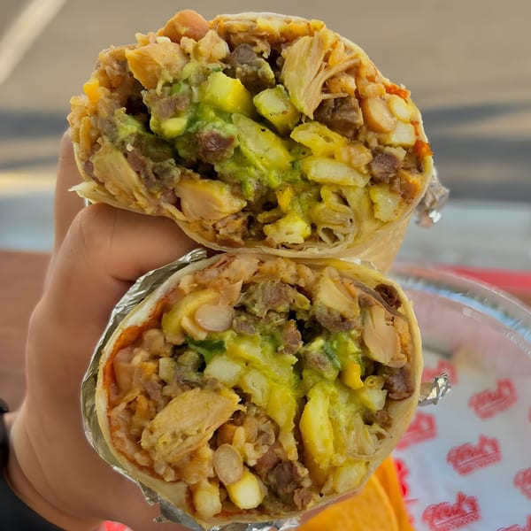 A hand holds a Fred's Red Tacos burrito, cut in half, revealing meat, fries, and guacamole.
