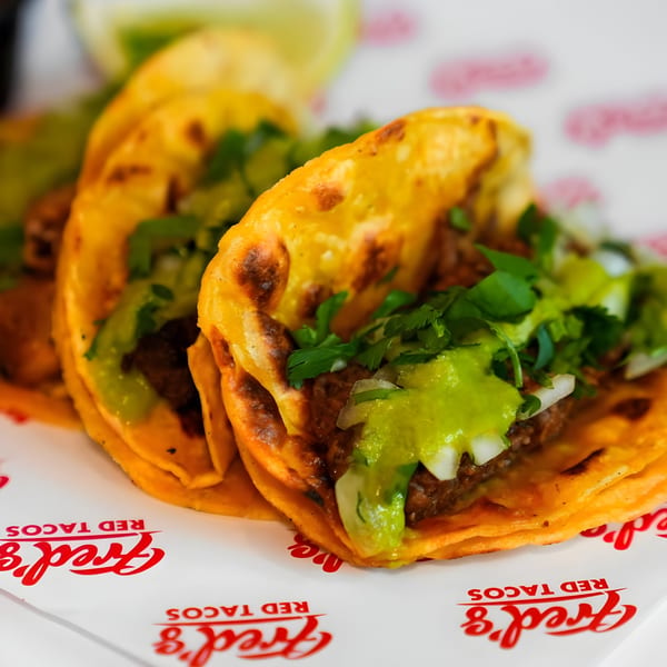 Three Fred's Red Tacos with green salsa, onions, and cilantro on a branded paper liner.
